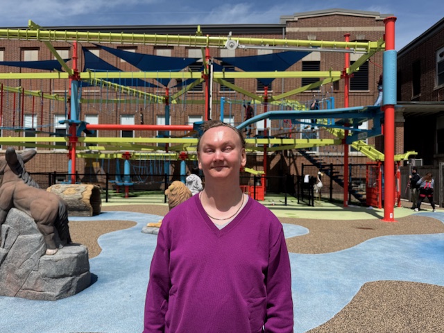 Cody Hurst, smiling in a purple sweater, standing outside the Missouri School for the Blind in front of its colorful outdoor ropes course.